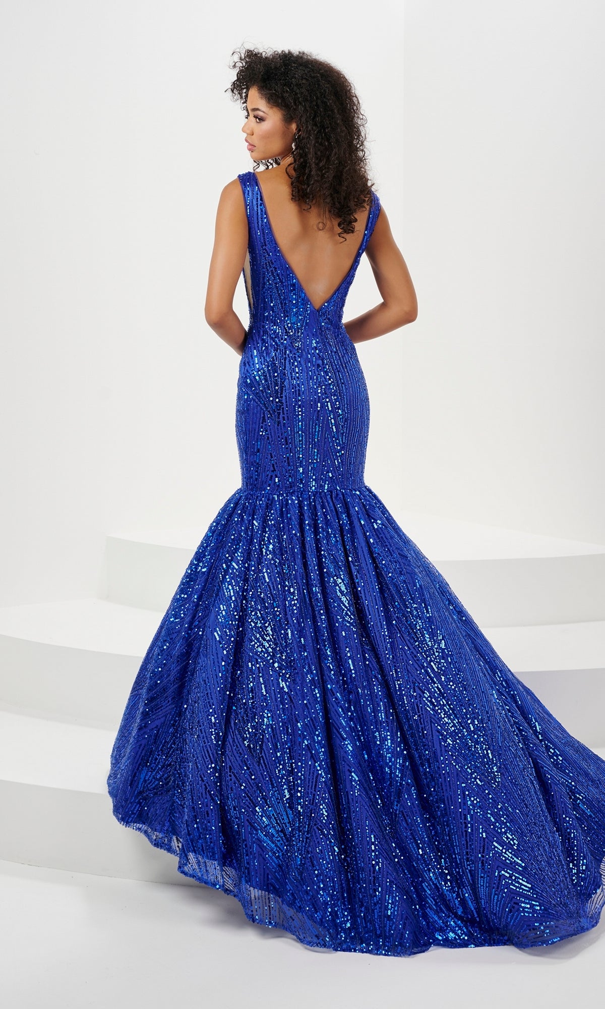 A woman in the Long Prom Dress 14180 by Panoply, a royal blue sleeveless sequin mermaid gown with a deep V-back and open back, stands on white steps facing away, showcasing its floor-length sparkling design.